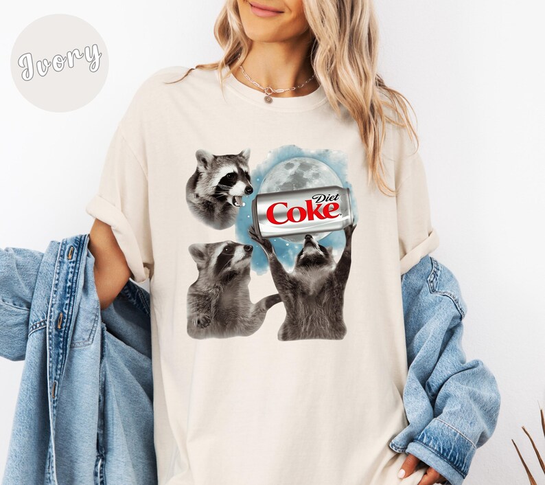 May include: Ivory-colored t-shirt featuring a graphic of three raccoons. One raccoon is holding a Diet Coke can in front of a moon, while the others look on. The text "Diet Coke" is visible on the can.