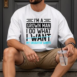 May include: White t-shirt with the text "I'M A GROWN MAN I DO WHAT I WANT MY DAUGHTER WANTS" in black and light blue. The shirt is worn by a person sitting outdoors.