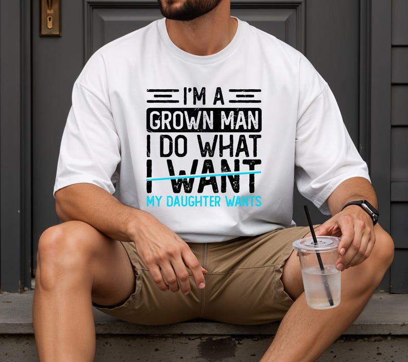 May include: White t-shirt with the text "I'M A GROWN MAN I DO WHAT I WANT MY DAUGHTER WANTS" in black and light blue. The shirt is worn by a person sitting outdoors.
