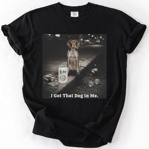 Pode incluir: Camiseta preta com um gráfico de um cão fumando um cigarro, uma lata de cerveja Lite e o texto "I Got That Dog In Me." O cão é da raça pointer marrom e branco.