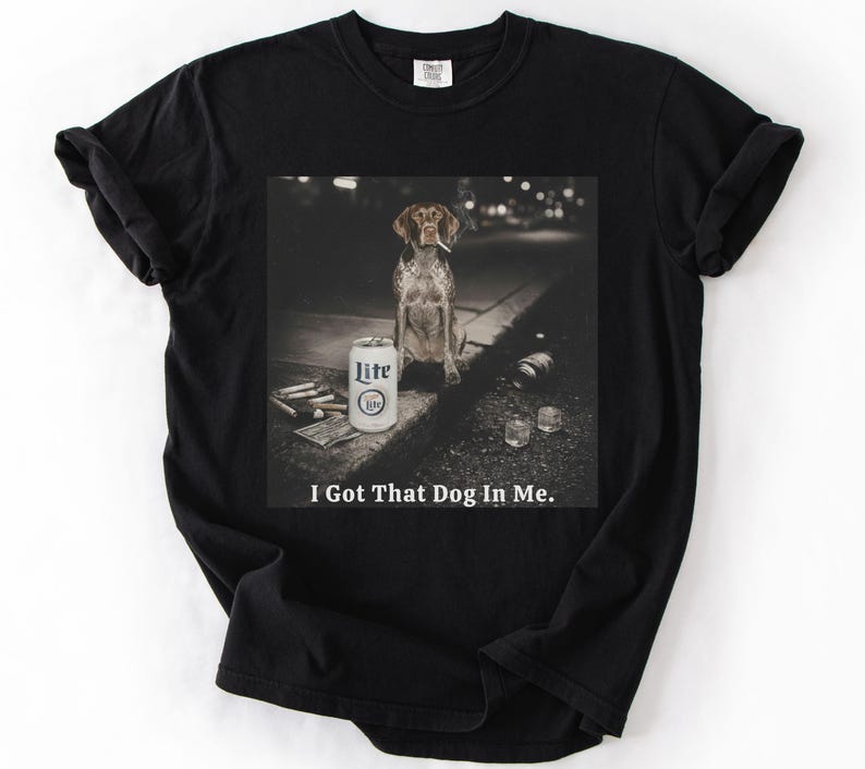 Pu&ograve; includere: T-shirt nero con una grafica di un cane che fuma una sigaretta, una lattina di birra Lite e la scritta "I Got That Dog In Me." Il cane &egrave; di razza pointer marrone e bianco.
