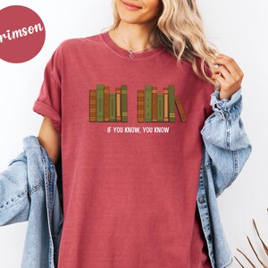 May include: Crimson-colored t-shirt featuring a graphic of a row of books in shades of green and brown. The text "IF YOU KNOW, YOU KNOW" is printed below the books. The shirt is a casual style.