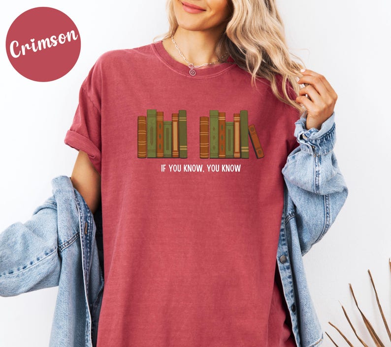 May include: Crimson-colored t-shirt featuring a graphic of a row of books in shades of green and brown. The text "IF YOU KNOW, YOU KNOW" is printed below the books. The shirt is a casual style.