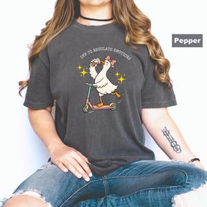 May include: Dark gray t-shirt featuring a cartoon goose riding a scooter, wearing a bow, and holding a drink. The text "OFF TO REGULATE EMOTIONS" is above the goose. The shirt is a relaxed fit.