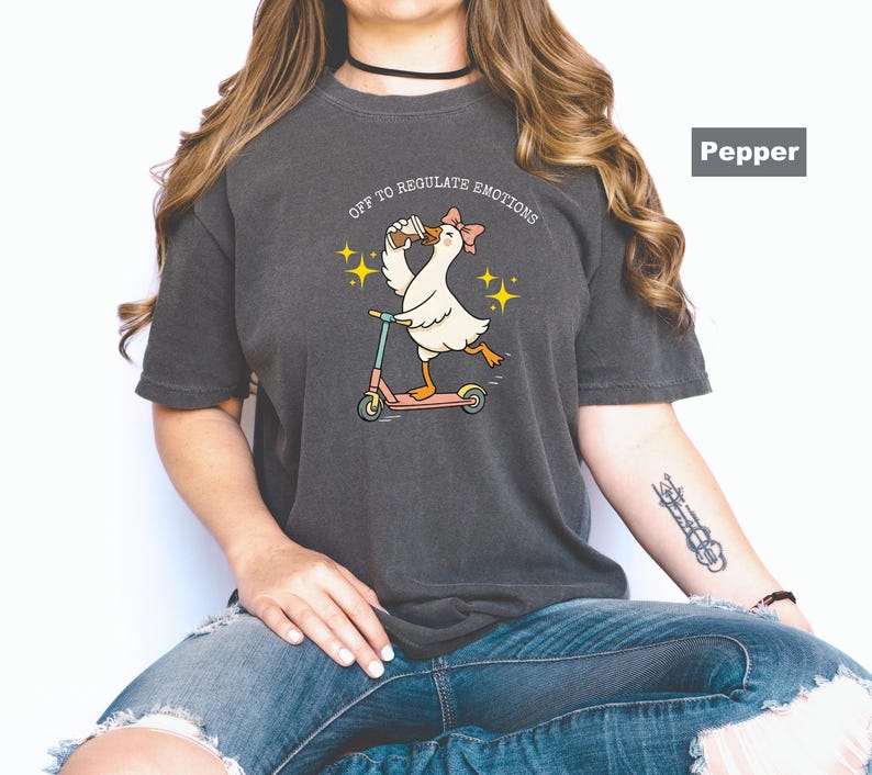 May include: Dark gray t-shirt featuring a cartoon goose riding a scooter, wearing a bow, and holding a drink. The text "OFF TO REGULATE EMOTIONS" is above the goose. The shirt is a relaxed fit.