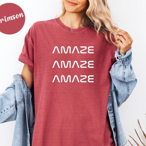 Pu&ograve; includere: T-shirt color cremisi con la parola "AMAZE" stampata tre volte in bianco. La maglietta ha maniche corte e una vestibilit&agrave; ampia. Una giacca di jeans &egrave; indossata sulle spalle.