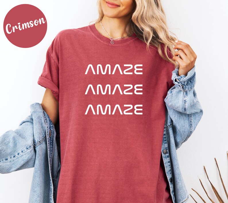 Pu&ograve; includere: T-shirt color cremisi con la parola "AMAZE" stampata tre volte in bianco. La maglietta ha maniche corte e una vestibilit&agrave; ampia. Una giacca di jeans &egrave; indossata sulle spalle.