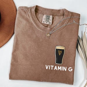 May include: A brown Comfort Colors t-shirt with a graphic of a pint of stout beer and the words "VITAMIN G" in white. A silver necklace with a pendant is draped across the shirt.