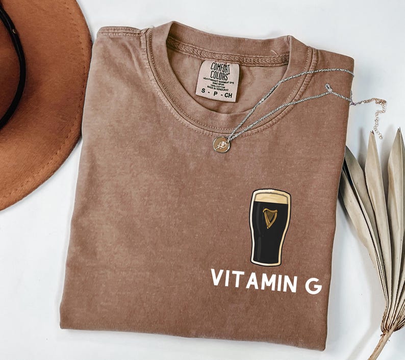 May include: A brown Comfort Colors t-shirt with a graphic of a pint of stout beer and the words "VITAMIN G" in white. A silver necklace with a pendant is draped across the shirt.