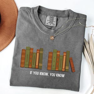 May include: Gray t-shirt with a graphic of stacked books in shades of green, orange, and brown. The text "IF YOU KNOW, YOU KNOW" is printed below the books. A silver necklace with a pendant is draped on the shirt.