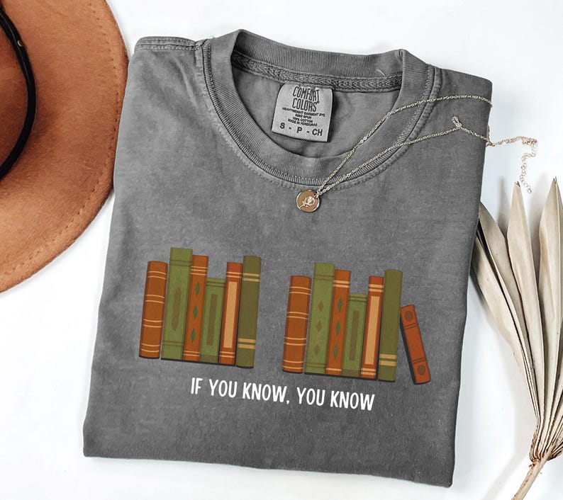 May include: Gray t-shirt with a graphic of stacked books in shades of green, orange, and brown. The text "IF YOU KNOW, YOU KNOW" is printed below the books. A silver necklace with a pendant is draped on the shirt.