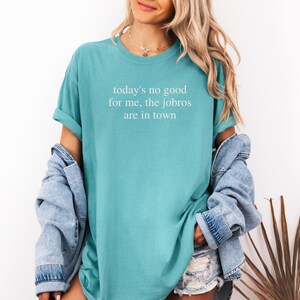 Op de afbeelding: Een turquoise t-shirt met de witte tekst "today's no good for me, the jobros are in town". Het shirt heeft korte mouwen en een relaxte pasvorm. Een spijkerjasje is over de schouders gedrapeerd.