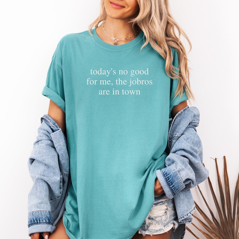 Op de afbeelding: Een turquoise t-shirt met de witte tekst "today's no good for me, the jobros are in town". Het shirt heeft korte mouwen en een relaxte pasvorm. Een spijkerjasje is over de schouders gedrapeerd.