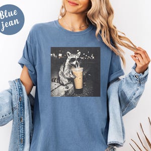 May include: Blue jean t-shirt with a graphic of a raccoon wearing sunglasses and drinking from a coffee cup. The raccoon is in a black and white image. The words "Blue jean" are in a blue circle in the upper left corner.