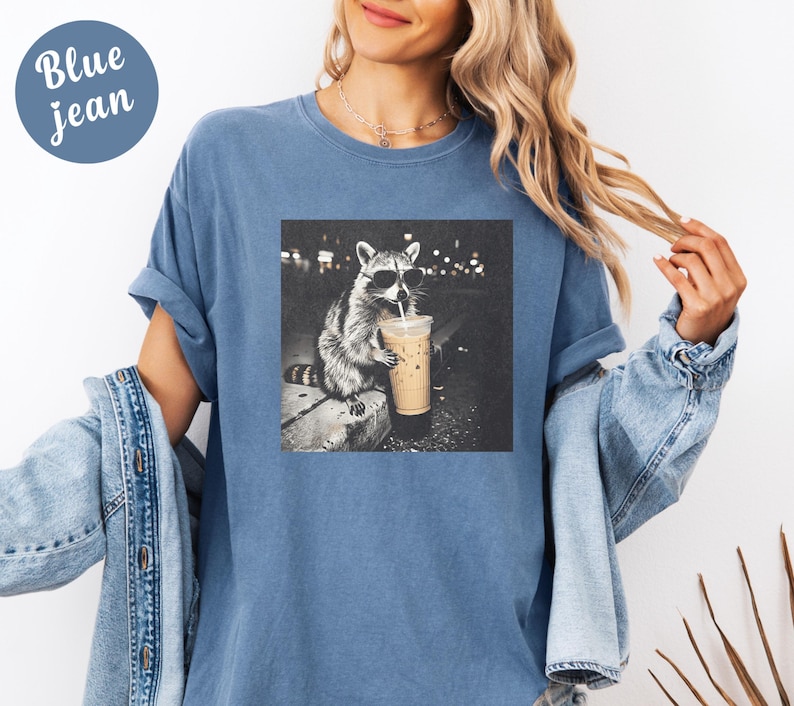 May include: Blue jean t-shirt with a graphic of a raccoon wearing sunglasses and drinking from a coffee cup. The raccoon is in a black and white image. The words "Blue jean" are in a blue circle in the upper left corner.
