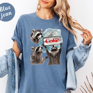 May include: A light blue t-shirt with a graphic of three raccoons and a Diet Coke can. The text "Diet Coke" is visible on the can. The shirt has short sleeves and a round neckline. The words "Blue jean" are in a blue circle in the upper left corner.