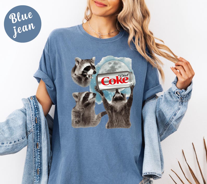 May include: A light blue t-shirt with a graphic of three raccoons and a Diet Coke can. The text "Diet Coke" is visible on the can. The shirt has short sleeves and a round neckline. The words "Blue jean" are in a blue circle in the upper left corner.