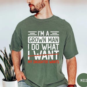 May include: Moss green t-shirt with white text that reads "I'M A GROWN MAN I DO WHAT I WANT" with "WANT" crossed out and replaced with "MY DAUGHTER WANTS" in red. The shirt is short-sleeved.