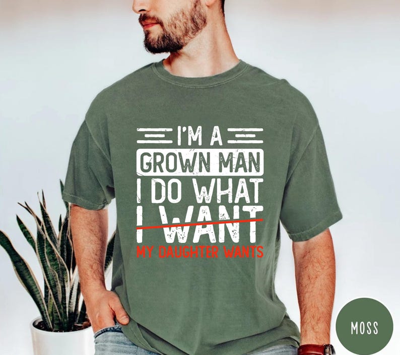 May include: Moss green t-shirt with white text that reads "I'M A GROWN MAN I DO WHAT I WANT" with "WANT" crossed out and replaced with "MY DAUGHTER WANTS" in red. The shirt is short-sleeved.
