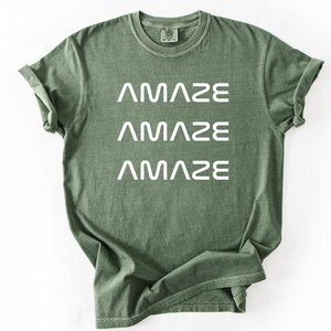 Pu&ograve; includere: T-shirt verde oliva con la scritta "AMAZE" stampata tre volte in bianco. La maglietta ha un girocollo e maniche corte. Il tessuto appare morbido e leggermente sbiadito.