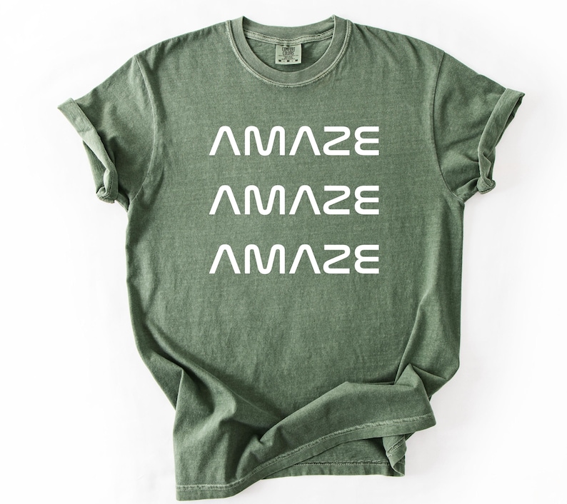 Pu&ograve; includere: T-shirt verde oliva con la scritta "AMAZE" stampata tre volte in bianco. La maglietta ha un girocollo e maniche corte. Il tessuto appare morbido e leggermente sbiadito.