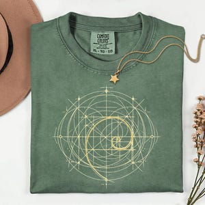 以下が含まれることがあります： セージグリーンのComfort Colors Tシャツ、ゴールドの幾何学模様。ゴールドの星型ペンダントネックレスがシャツの上にあります。シャツには「Comfort Colors」とサイズ「XL」と書かれたタグが付いています。
