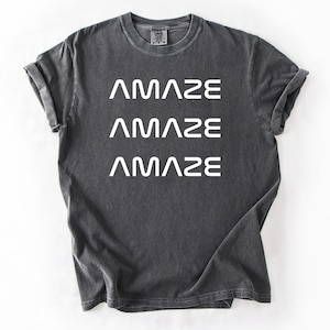 Pu&ograve; includere: T-shirt grigio scuro con la parola "AMAZE" stampata tre volte in bianco, impilata verticalmente. La maglietta ha un girocollo e maniche arrotolate. Il tessuto sembra prelavato per un aspetto vintage.
