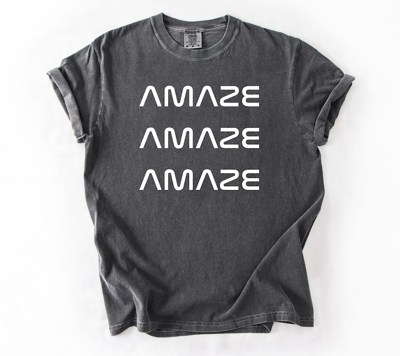 Pu&ograve; includere: T-shirt grigio scuro con la parola "AMAZE" stampata tre volte in bianco, impilata verticalmente. La maglietta ha un girocollo e maniche arrotolate. Il tessuto sembra prelavato per un aspetto vintage.