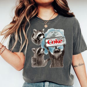 May include: Dark gray t-shirt featuring a graphic of three raccoons and a Diet Coke can. The raccoons are in various poses, with one holding the can towards a moon graphic. The Diet Coke can is red and white.