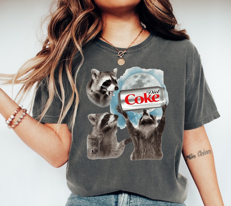 May include: Dark gray t-shirt featuring a graphic of three raccoons and a Diet Coke can. The raccoons are in various poses, with one holding the can towards a moon graphic. The Diet Coke can is red and white.