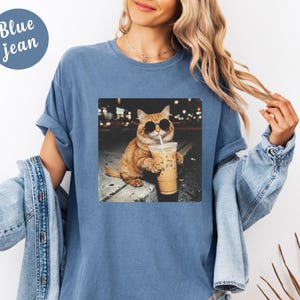 May include: Blue jean t-shirt with a graphic of a cat wearing sunglasses and drinking iced coffee. The cat is orange and white, and the image has a nighttime city background. The t-shirt has short sleeves and a round neckline.