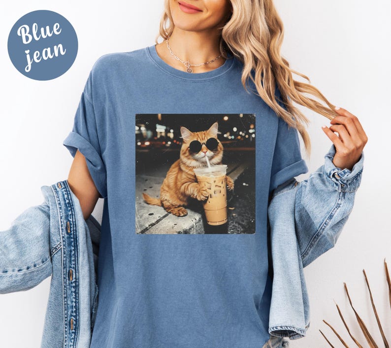 May include: Blue jean t-shirt with a graphic of a cat wearing sunglasses and drinking iced coffee. The cat is orange and white, and the image has a nighttime city background. The t-shirt has short sleeves and a round neckline.