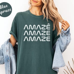 May include: A blue spruce-colored t-shirt with the word "AMAZE" stacked three times in white, with a small graphic above the last "E". The shirt has a relaxed fit and rolled sleeves.