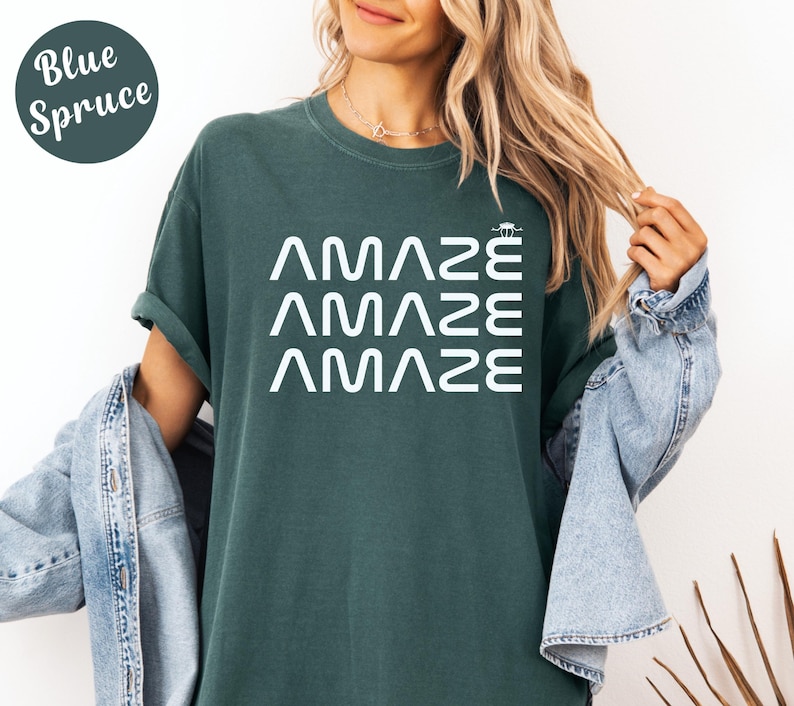 May include: A blue spruce-colored t-shirt with the word "AMAZE" stacked three times in white, with a small graphic above the last "E". The shirt has a relaxed fit and rolled sleeves.