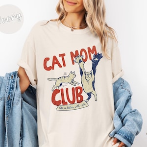 May include: Ivory-colored t-shirt with a retro design featuring the text "CAT MOM CLUB" in red and blue. The graphic includes two cartoon cats, one jumping and the other standing upright. The phrase "life is better with cats" is also included.