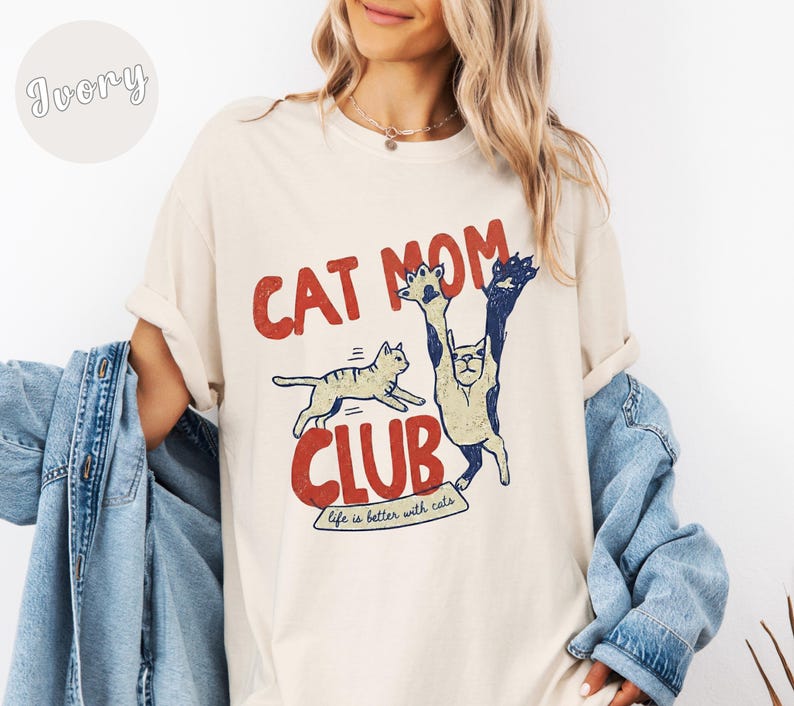May include: Ivory-colored t-shirt with a retro design featuring the text "CAT MOM CLUB" in red and blue. The graphic includes two cartoon cats, one jumping and the other standing upright. The phrase "life is better with cats" is also included.