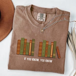 May include: A brown t-shirt with a graphic of stacked books in shades of green and orange. The text "IF YOU KNOW, YOU KNOW" is printed below the books. A silver necklace with a pendant is on the shirt.