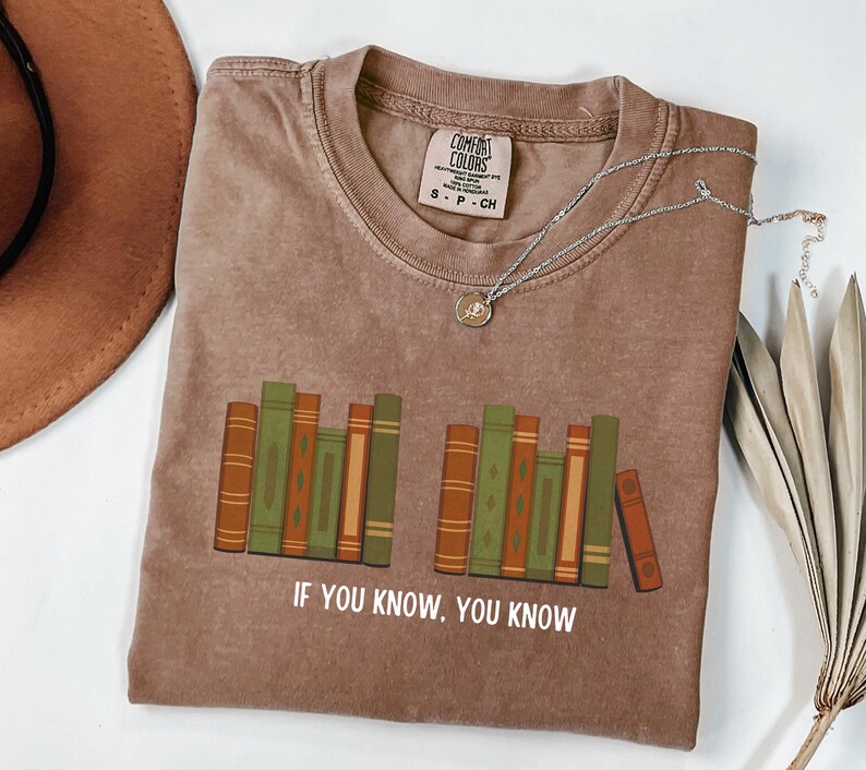 May include: A brown t-shirt with a graphic of stacked books in shades of green and orange. The text "IF YOU KNOW, YOU KNOW" is printed below the books. A silver necklace with a pendant is on the shirt.