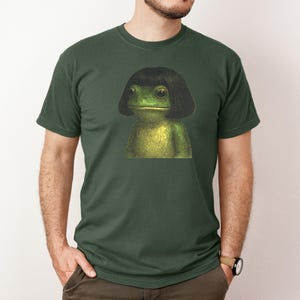 May include: Dark green t-shirt featuring a whimsical illustration of a frog with a black bob hairstyle. The frog is depicted in shades of green and yellow, creating a unique and eye-catching design. The t-shirt is a classic crew neck style.
