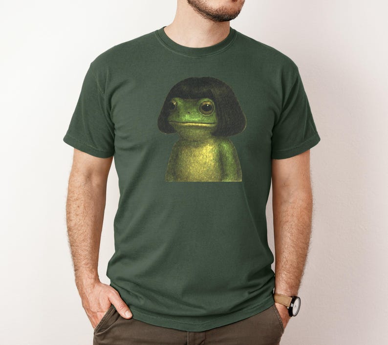 May include: Dark green t-shirt featuring a whimsical illustration of a frog with a black bob hairstyle. The frog is depicted in shades of green and yellow, creating a unique and eye-catching design. The t-shirt is a classic crew neck style.