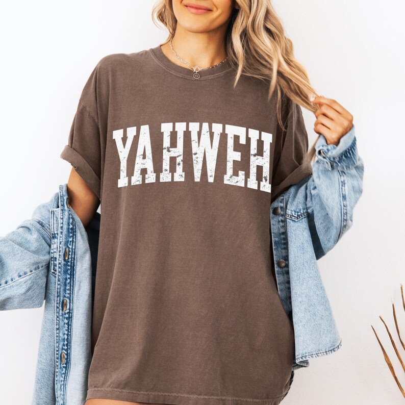 May include: A brown t-shirt with the word "YAHWEH" printed in white, distressed lettering. The shirt has short sleeves and a relaxed fit. A denim jacket is draped over the shoulders.