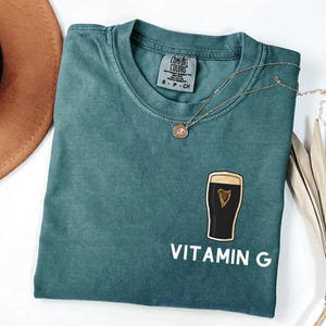 May include: A teal Comfort Colors t-shirt with a graphic of a pint of stout and the words "VITAMIN G" in white. A gold necklace with a small pendant rests on the shirt. The shirt is folded and displayed on a white surface.