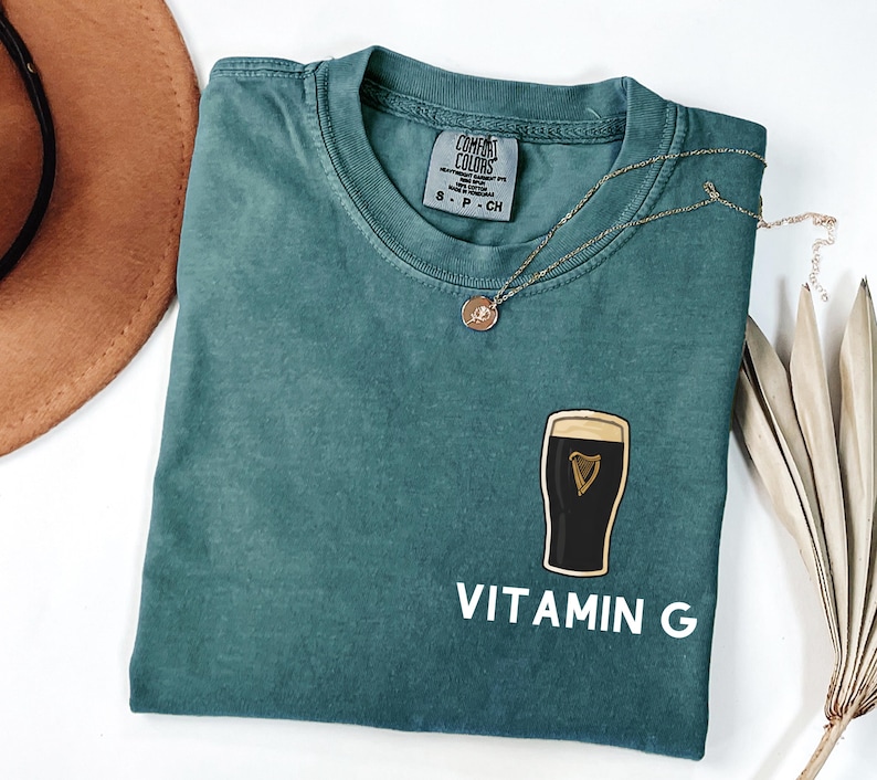 May include: A teal Comfort Colors t-shirt with a graphic of a pint of stout and the words "VITAMIN G" in white. A gold necklace with a small pendant rests on the shirt. The shirt is folded and displayed on a white surface.