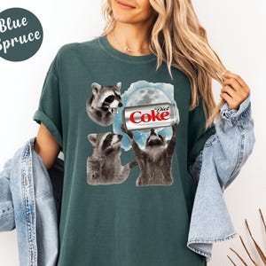 May include: Blue Spruce t-shirt featuring a graphic of three raccoons and a Diet Coke can. The raccoons are depicted in a cartoon style, with one holding the can towards a full moon. The shirt is a solid color.