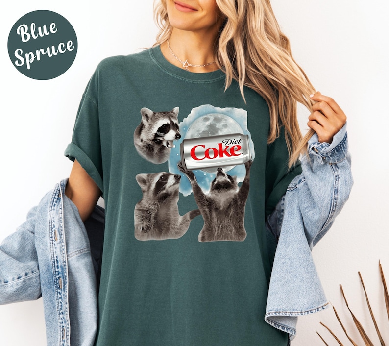 May include: Blue Spruce t-shirt featuring a graphic of three raccoons and a Diet Coke can. The raccoons are depicted in a cartoon style, with one holding the can towards a full moon. The shirt is a solid color.