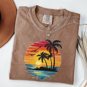 May include: A brown Comfort Colors t-shirt featuring an embroidered sunset scene with palm trees, birds, and ocean waves. A silver necklace with a pendant rests on the shirt. A brown hat and dried leaves are also in the image.