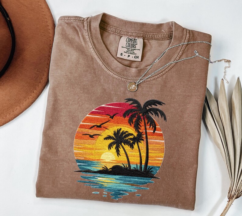 May include: A brown Comfort Colors t-shirt featuring an embroidered sunset scene with palm trees, birds, and ocean waves. A silver necklace with a pendant rests on the shirt. A brown hat and dried leaves are also in the image.