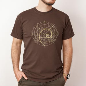 以下が含まれることがあります： 黄金のスパイラルデザインが特徴の茶色のTシャツ。グラフィックは線と円の幾何学模様です。シャツはクルーネックで半袖です。その人は茶色のストラップの時計をしています。