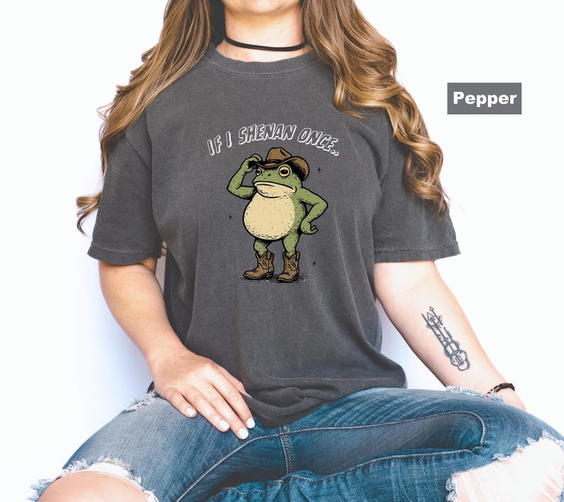 May include: Dark grey t-shirt with a cartoon frog in a cowboy hat and boots. The text "IF I SHENAN ONCE..." is above the frog. The shirt has a relaxed fit.