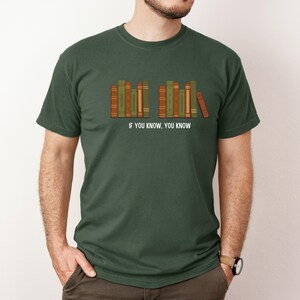 May include: Dark green t-shirt featuring a row of stylized books in shades of green, orange, and brown. The text "IF YOU KNOW. YOU KNOW" is printed below the books. The shirt is a crew neck style.